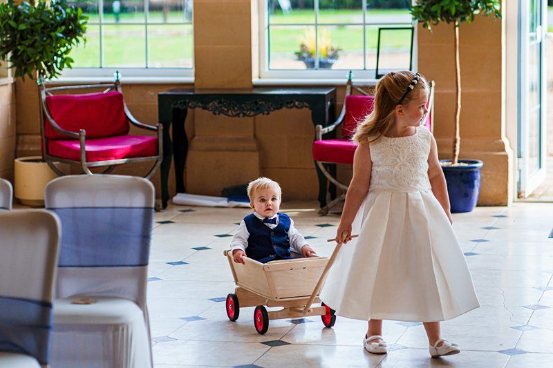 A young girl in a white dress is pulling a wooden wagon with a small boy in a blue vest and white shirt sitting inside. They are indoors with large windows, potted plants, and red chairs in the background. The girl and boy appear to be part of a formal event. Image by Documentary Wedding Photographer Lewis Bishop Photography.