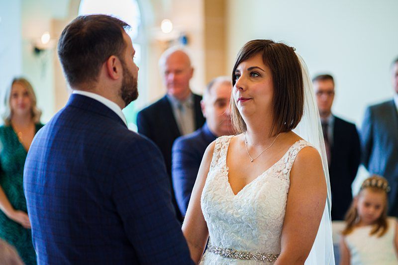 A bride and groom stand facing each other holding hands during their wedding ceremony. The bride is in a white lace gown with a veil, and the groom is in a dark suit. Guests are seated and standing in the background, watching the ceremony. Image by Documentary Wedding Photographer Lewis Bishop Photography.