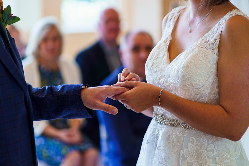 Close-up image of a bride and groom during their wedding ceremony. The bride, wearing a white lace gown, is placing a ring on the groom's finger. Several guests can be seen in the softly focused background, seated and watching the moment. Image by Documentary Wedding Photographer Lewis Bishop Photography.