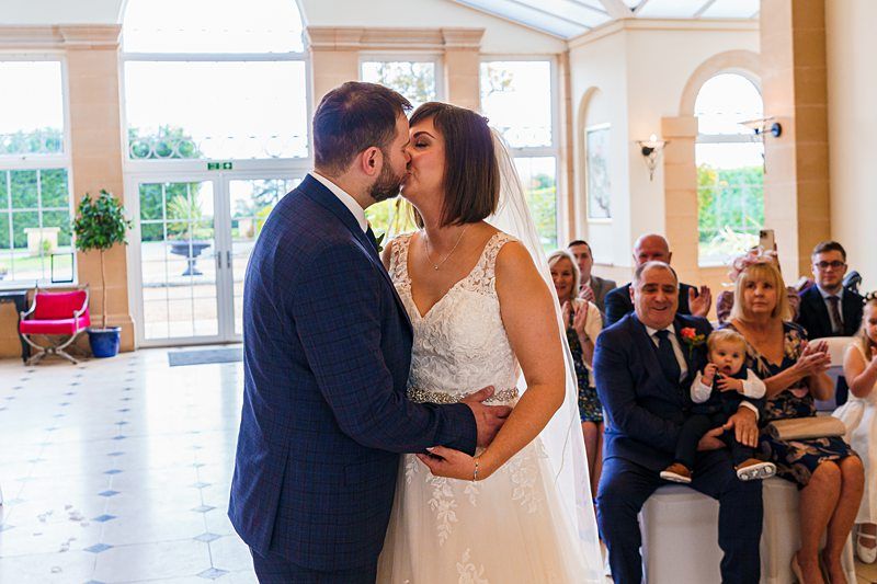 A bride and groom share a kiss during their wedding ceremony in a bright, sunlit room. The bride wears a white gown and veil, while the groom wears a dark suit. Seated guests, some clapping and one holding a baby, look on and smile. Image by Documentary Wedding Photographer Lewis Bishop Photography.