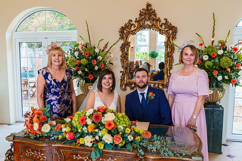 A newlywed couple sit at an ornate table in front of a large mirror, with the bride in a white dress and the groom in a blue suit. Two women stand on either side, all smiling. The table is adorned with vibrant flowers, and floral arrangements decorate the background. Image by Documentary Wedding Photographer Lewis Bishop Photography.