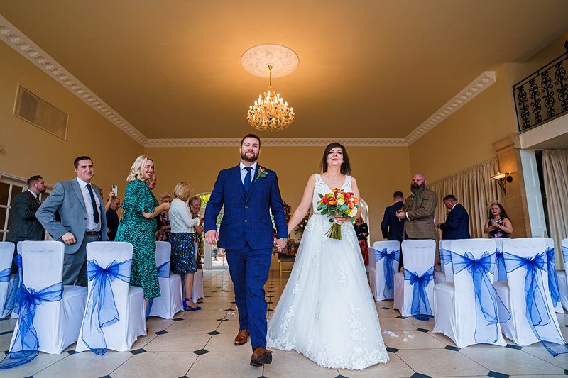 A newly married couple, the bride in a white gown holding a bouquet of flowers and the groom in a blue suit, walk down the aisle of a decorated wedding venue. Guests on either side, seated on chairs adorned with blue ribbons, applaud them. A chandelier hangs overhead. Image by Documentary Wedding Photographer Lewis Bishop Photography.