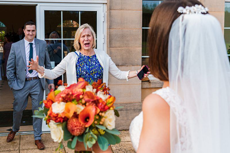 A bride in a white dress and veil holds a bouquet of orange flowers. A woman in a blue dress and white cardigan stands with her arms wide open, excitedly greeting the bride. A man in a suit stands in the background, smiling. They are outside a building with large windows. Image by Documentary Wedding Photographer Lewis Bishop Photography.