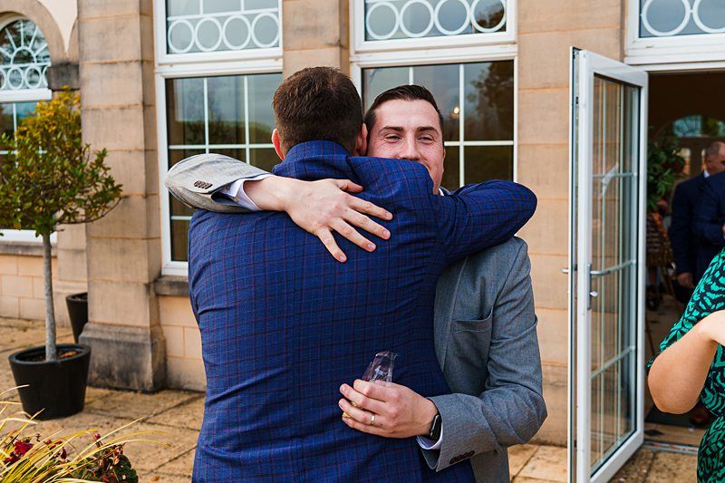 Two men hugging outside a building with large windows and an open glass door. One man, in a blue checkered suit, has his back to the camera, while the other, wearing a gray suit, faces forward and smiles. There are other people and greenery in the background. Image by Documentary Wedding Photographer Lewis Bishop Photography.