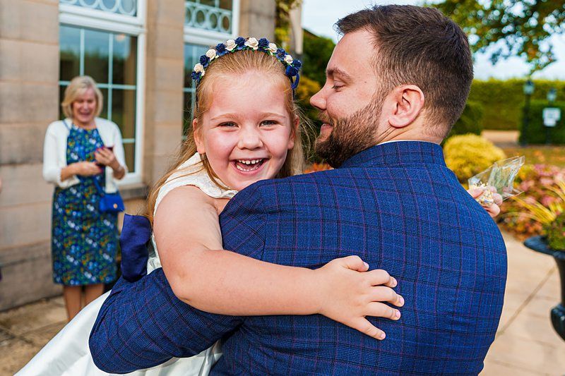 A man in a blue checkered suit holds a smiling young girl wearing a white dress and a flower headband. They are outdoors in front of a building with large windows. A woman in the background claps and smiles. Image by Documentary Wedding Photographer Lewis Bishop Photography.