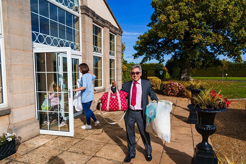 A man in a suit and tie smiles while carrying a large white bag, a red polka dot bag, and a blue bag. A woman and a child enter a large, glass-door building next to him. The setting is outdoors with trees and plants in the background. Image by Documentary Wedding Photographer Lewis Bishop Photography.