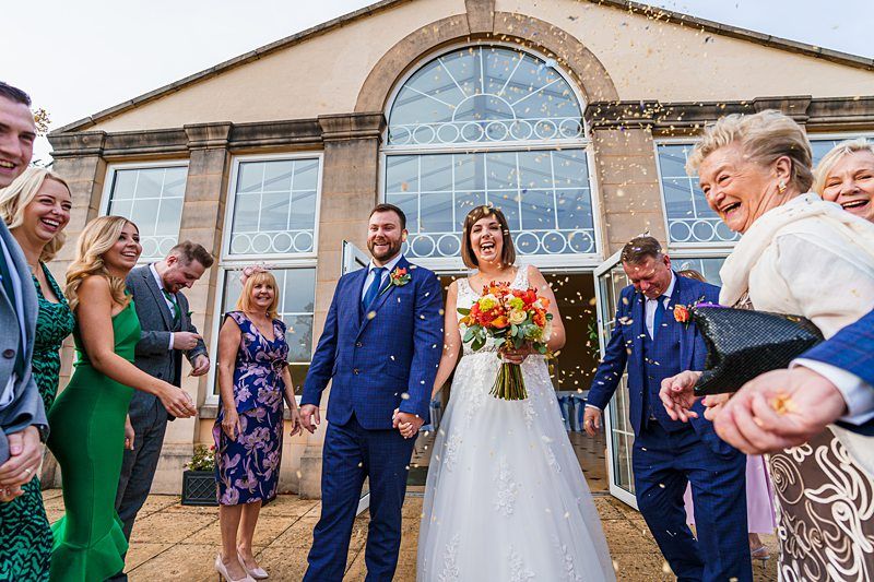 A joyous wedding scene outside a large, elegant building with big windows. The bride, in a white dress holding a bouquet, and the groom, in a blue suit, smile as guests cheer and throw confetti. Everyone looks happy and festive. Image by Documentary Wedding Photographer Lewis Bishop Photography.