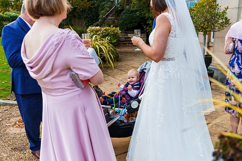 A bride in a white dress and veil stands outdoors beside a stroller holding a smiling baby. Another woman in a pink dress faces the baby, holding a drink. Part of another person's blue suit is visible on the left. Greenery and stone steps are in the background. Image by Documentary Wedding Photographer Lewis Bishop Photography.