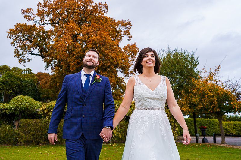 A happy couple, dressed in wedding attire, walks hand in hand through a park with vibrant autumn trees. The groom is in a blue suit, and the bride is in a white gown, both smiling as they enjoy the outdoor setting. Image by Documentary Wedding Photographer Lewis Bishop Photography.