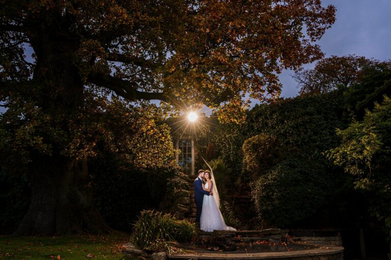 A bride and groom stand together under a large tree with autumn-colored leaves. The couple is illuminated by a bright light behind them, creating a glowing effect. They are surrounded by lush vegetation and an old stone structure partially covered with ivy. Image by Documentary Wedding Photographer Lewis Bishop Photography.