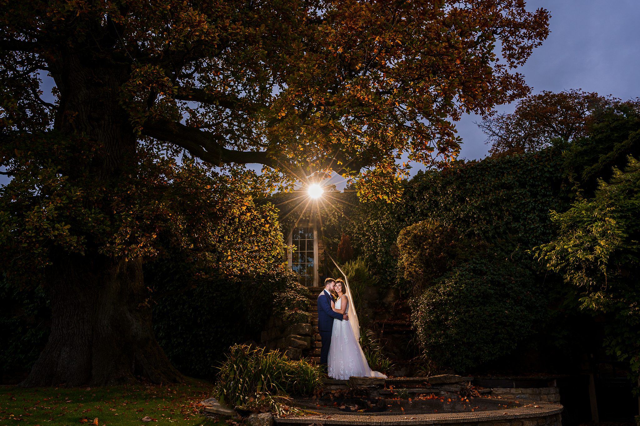 A bride and groom stand together under a large tree with autumn-colored leaves. The couple is illuminated by a bright light behind them, creating a glowing effect. They are surrounded by lush vegetation and an old stone structure partially covered with ivy. Image by Documentary Wedding Photographer Lewis Bishop Photography.