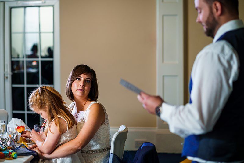 A woman with a short bob haircut sits at a table holding a young girl in her lap. The girl is focused on something in front of her. A man in a white shirt and blue waistcoat stands nearby, holding a piece of paper and presumably giving a speech. Image by Documentary Wedding Photographer Lewis Bishop Photography.