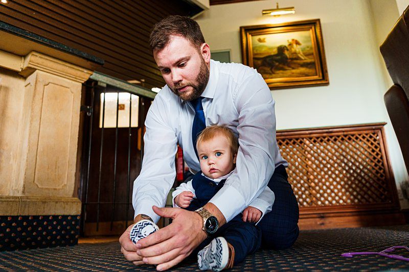 A man wearing a white shirt and tie helps a baby put on their socks. The baby, dressed in a blue outfit, sits calmly as the man, kneeling on the carpeted floor, focuses on the task. Behind them, a painting hangs on the wall in a warmly lit room. Image by Documentary Wedding Photographer Lewis Bishop Photography.