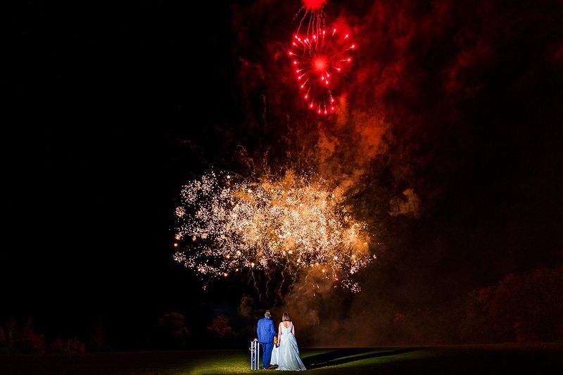 Two people, one in a blue outfit and the other in a white dress, stand on a grassy field at night, viewing a vibrant fireworks display. Red and golden fireworks illuminate the sky, creating a beautiful scene against the dark backdrop. Image by Documentary Wedding Photographer Lewis Bishop Photography.
