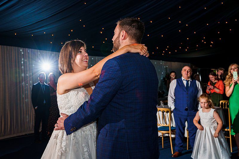 A bride and groom share a first dance at their wedding reception under a starry canopy of lights. The bride, in a white gown, smiles at the groom, who wears a blue suit. Guests, including a child in a white dress, watch and capture the moment. Image by Documentary Wedding Photographer Lewis Bishop Photography.