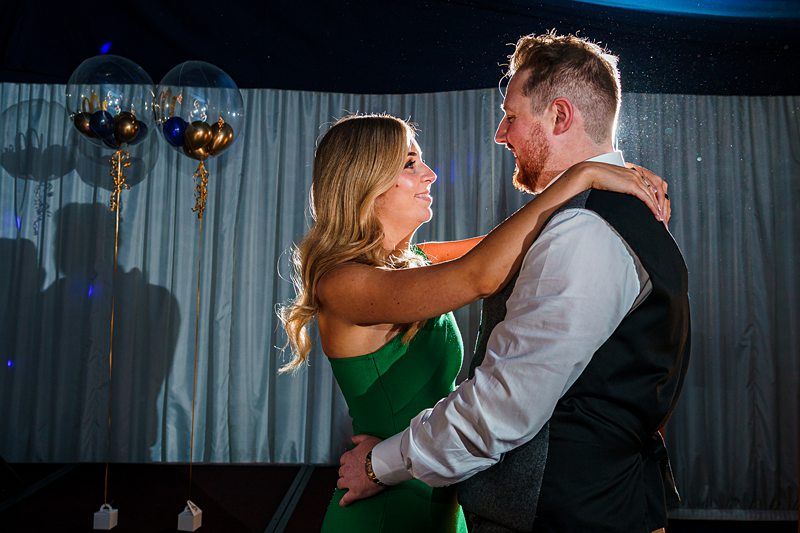 A couple embraces while dancing under a spotlight. The woman, wearing a green dress, and the man, in a white shirt and black vest, gaze into each other's eyes. In the background, there are gold and blue balloons and a curtain backdrop. Image by Documentary Wedding Photographer Lewis Bishop Photography.