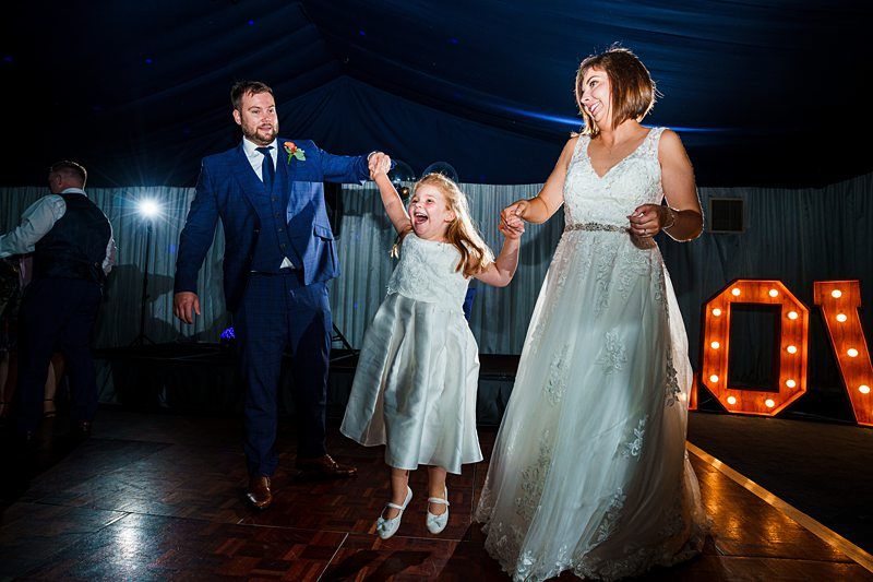 A man in a blue suit and a woman in a white bridal dress joyfully dance with a young girl in a white dress on a dance floor. Large illuminated letters spelling "LOVE" are in the background. The atmosphere is lively with colorful lighting. Image by Documentary Wedding Photographer Lewis Bishop Photography.