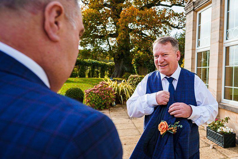 Two groomsmen in blue plaid vests are standing outside a building with large windows. One is smiling while holding his jacket, which has an orange rose boutonniere. A well-kept garden and autumn trees are in the background under a clear sky. Image by Documentary Wedding Photographer Lewis Bishop Photography.