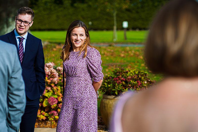 A woman in a purple dress with a floral pattern is smiling outdoors, standing near colorful flowering plants. A man in a suit and tie stands beside her. Both are engaged in conversation with another person whose back is to the camera. Image by Documentary Wedding Photographer Lewis Bishop Photography.