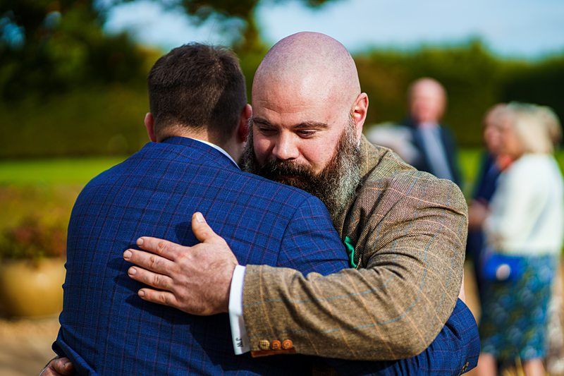 Two men are sharing an embrace outdoors, both wearing suits. The man facing the camera has a bald head and a full beard, while the other man's back is turned. Several blurred people are in the background, along with greenery and a bright blue sky. Image by Documentary Wedding Photographer Lewis Bishop Photography.