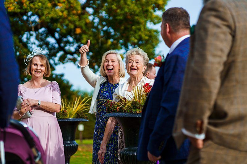 A group of people standing outside on a sunny day, with a green background. Three women are smiling and one is giving a thumbs-up. The women are dressed in formal attire, and two men are partially visible from behind, also dressed in suits. Image by Documentary Wedding Photographer Lewis Bishop Photography.