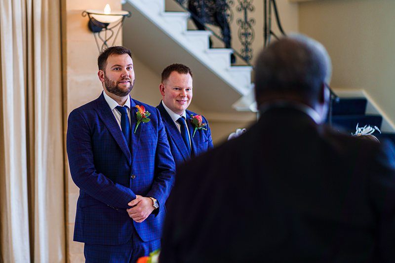 Two groomsmen in blue suits with orange boutonnieres stand smiling, facing an officiant with their backs to a staircase. The setting appears to be indoors, with light-colored curtains and a lit wall sconce in the background. Image by Documentary Wedding Photographer Lewis Bishop Photography.