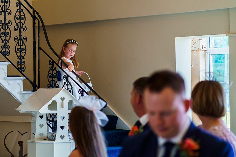A young girl with a flower crown stands on a staircase holding a basket, looking toward a group of adults below who are engaged in conversation. The staircase has an ornate black railing, and there is a small white house-shaped decoration nearby. Image by Documentary Wedding Photographer Lewis Bishop Photography.