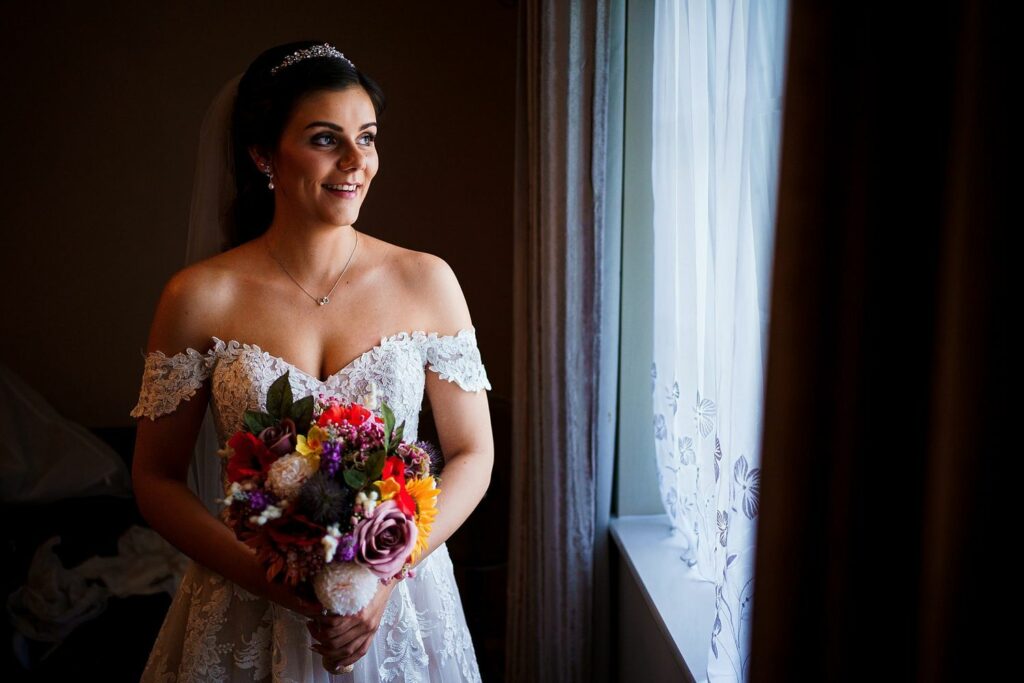 A bride wearing a white off-the-shoulder wedding dress and veil stands by a window, holding a colorful bouquet of flowers. She is smiling and looking outside, with soft light illuminating her face and dress from the window. Image by Documentary Wedding Photographer Lewis Bishop Photography.