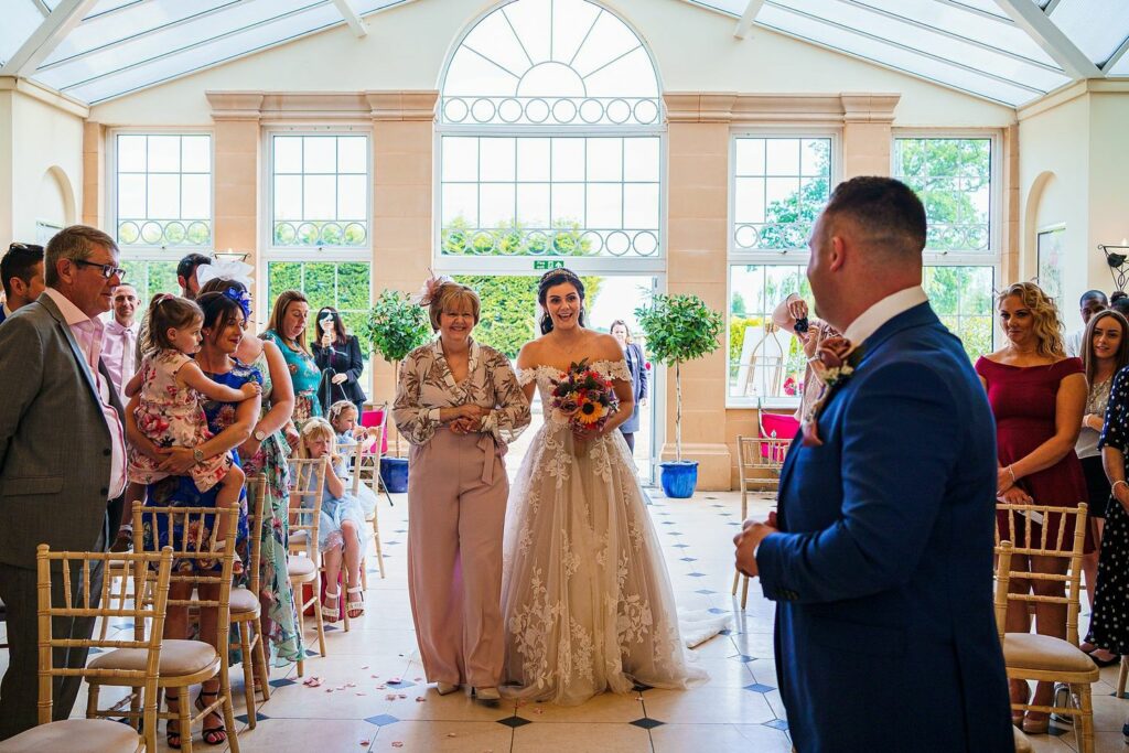 A bride in a white dress holds a bouquet and smiles as she walks down the aisle accompanied by an older woman. Guests stand on either side, watching, while a man in a blue suit, possibly the groom, stands at the front of the aisle in an elegant, sunlit venue. Image by Documentary Wedding Photographer Lewis Bishop Photography.