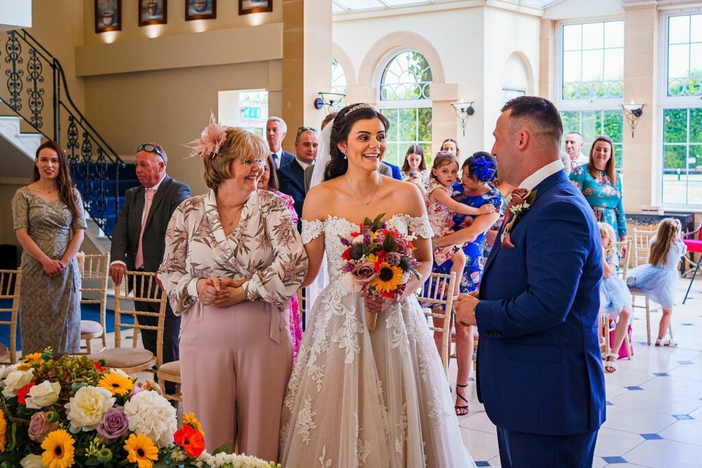 A bride in a white gown and veil, holding a colorful bouquet, stands smiling next to a woman in a floral blouse. A man in a blue suit stands facing them. Guests seated and standing in a brightly lit room, with large windows and flowers in the foreground. Image by Documentary Wedding Photographer Lewis Bishop Photography.