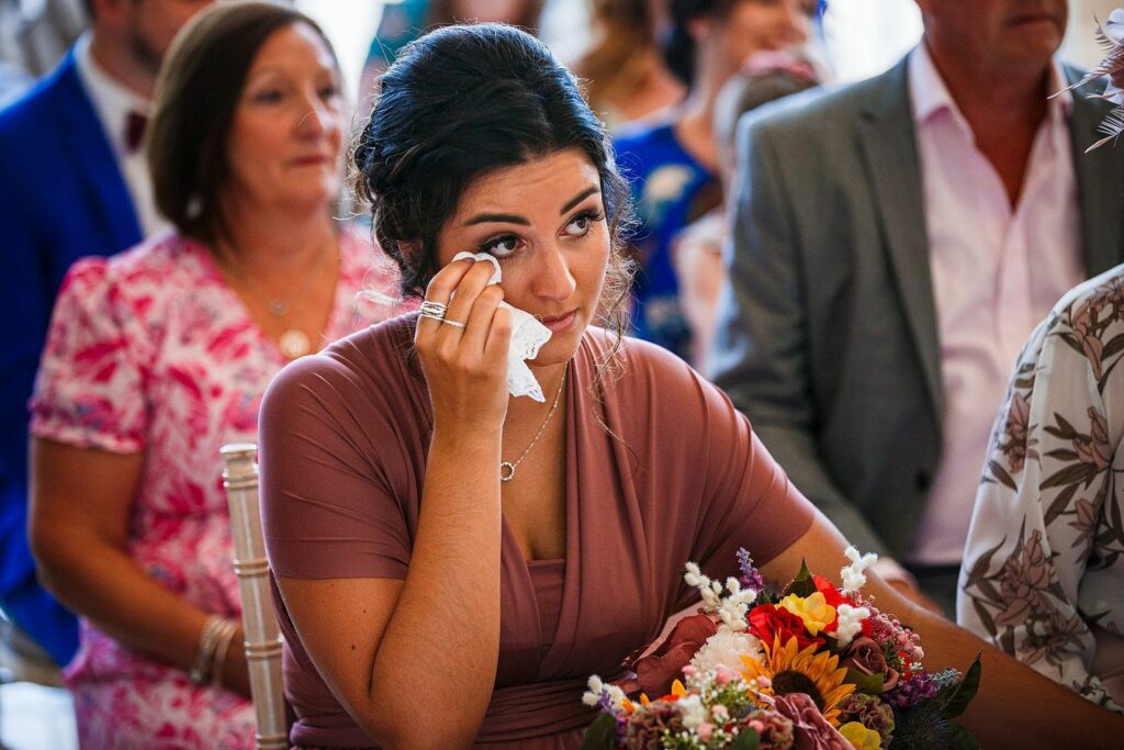 A woman in a mauve dress dabs her eyes with a tissue, appearing emotional during what seems to be a wedding ceremony. She is seated and holding a bouquet of colorful flowers. Several other guests, dressed in various formal attire, are seated around her. Image by Documentary Wedding Photographer Lewis Bishop Photography.