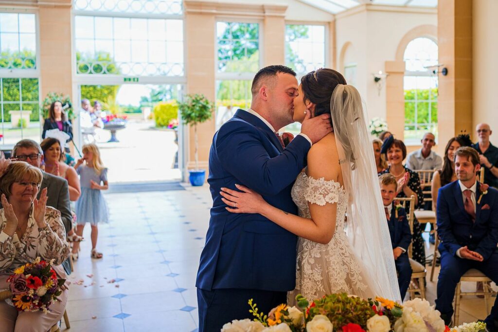 A newlywed couple shares a kiss at their wedding ceremony in a bright, elegant venue. The bride wears a white lace gown and veil, while the groom is in a blue suit. Guests, young and old, watch and clap, surrounded by flowers and sunlight streaming through large windows. Image by Documentary Wedding Photographer Lewis Bishop Photography.