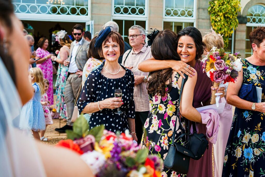 A group of people at an outdoor event, with a woman in a floral dress hugging another woman holding a bouquet. Others are smiling and chatting in the background. The scene is cheerful and festive, with colorful flowers and well-dressed individuals enjoying the occasion. Image by Documentary Wedding Photographer Lewis Bishop Photography.