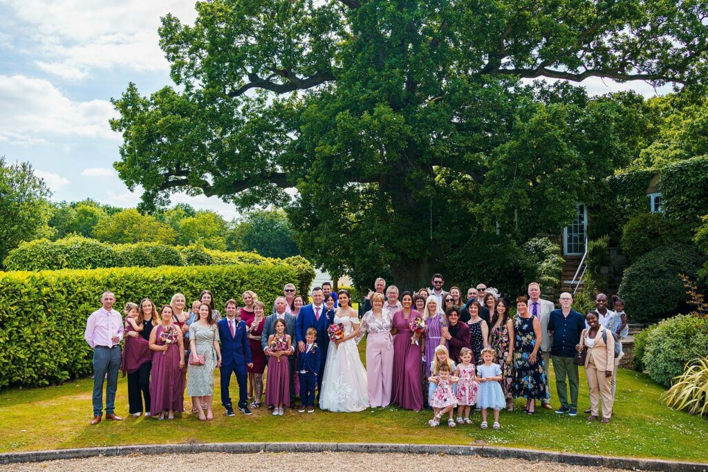 A large wedding party poses for a group photo outdoors. The bride and groom are at the center, surrounded by bridesmaids in purple dresses, groomsmen in suits, and guests dressed in various outfits. They stand in front of a large tree and lush greenery on a sunny day. Image by Documentary Wedding Photographer Lewis Bishop Photography.