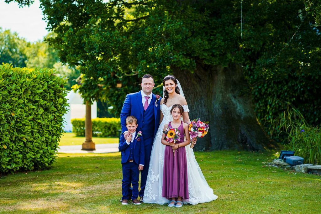 A wedding couple stands outdoors with a tree in the background. The bride, in a white gown and veil, holds a colorful bouquet. Beside her is the groom in a blue suit. In front are two children, a boy in a suit and a girl in a purple dress holding a bouquet. Image by Documentary Wedding Photographer Lewis Bishop Photography.
