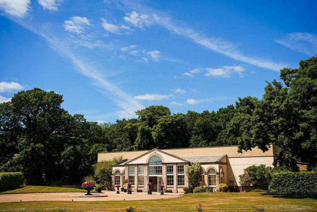 A picturesque, single-story villa framed by lush greenery, under a clear blue sky with wisps of clouds. The villa features large arched windows and a central arched entrance, surrounded by a well-manicured lawn and vibrant flower arrangements. Image by Documentary Wedding Photographer Lewis Bishop Photography.