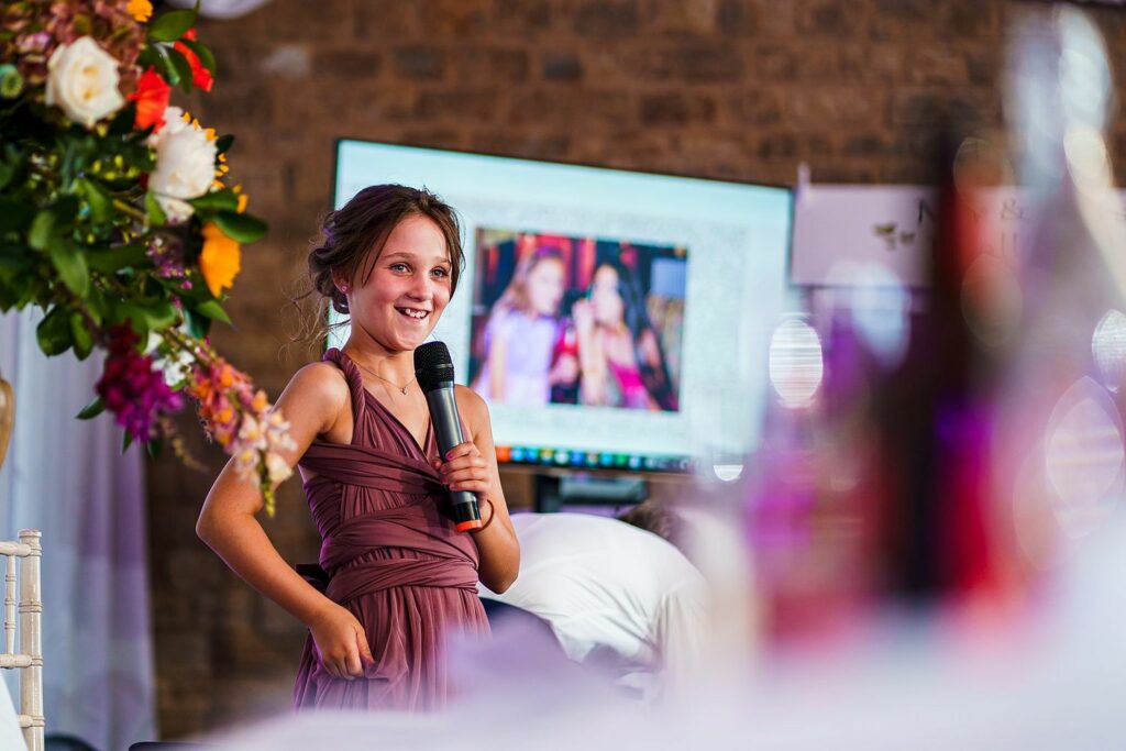 A young girl in a burgundy dress stands holding a microphone, smiling and looking to the side. She is positioned in front of a large display screen showing a blurred image. Bright flowers and soft decorations are visible in the foreground. Image by Documentary Wedding Photographer Lewis Bishop Photography.