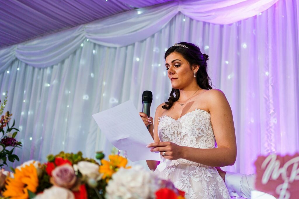 A bride in a white, strapless gown with lace details is standing and speaking into a microphone while holding a piece of paper. Behind her is a draped, white curtain with soft, purple lighting. In the foreground, colorful flowers are visible. Image by Documentary Wedding Photographer Lewis Bishop Photography.
