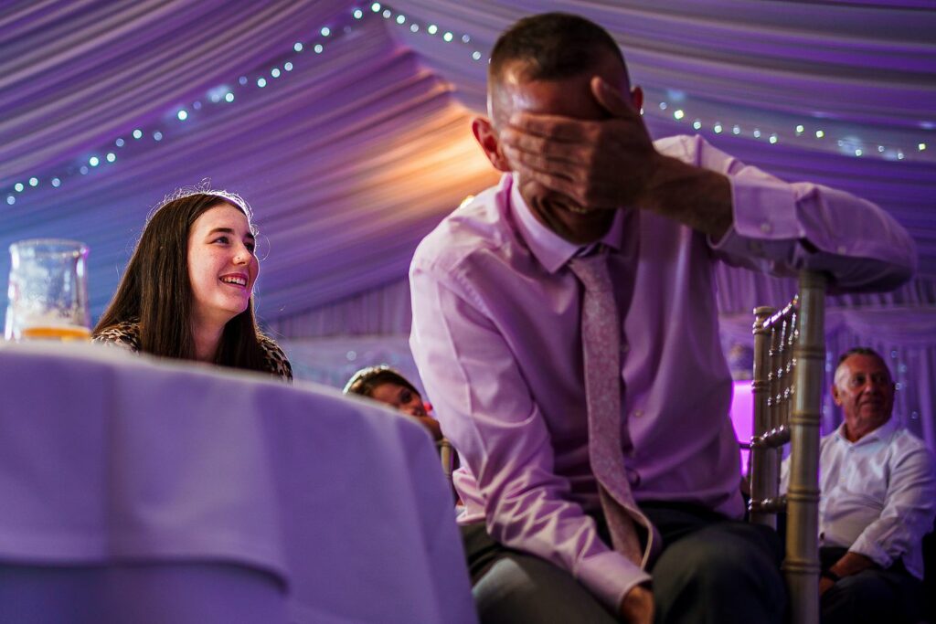 A man in a pink shirt and tie sits on a chair, covering his face with one hand in apparent laughter. A woman with long dark hair, sitting next to him, looks on and smiles. They are in a brightly lit room, decorated with string lights on the ceiling. Other people are visible in the background. Image by Documentary Wedding Photographer Lewis Bishop Photography.