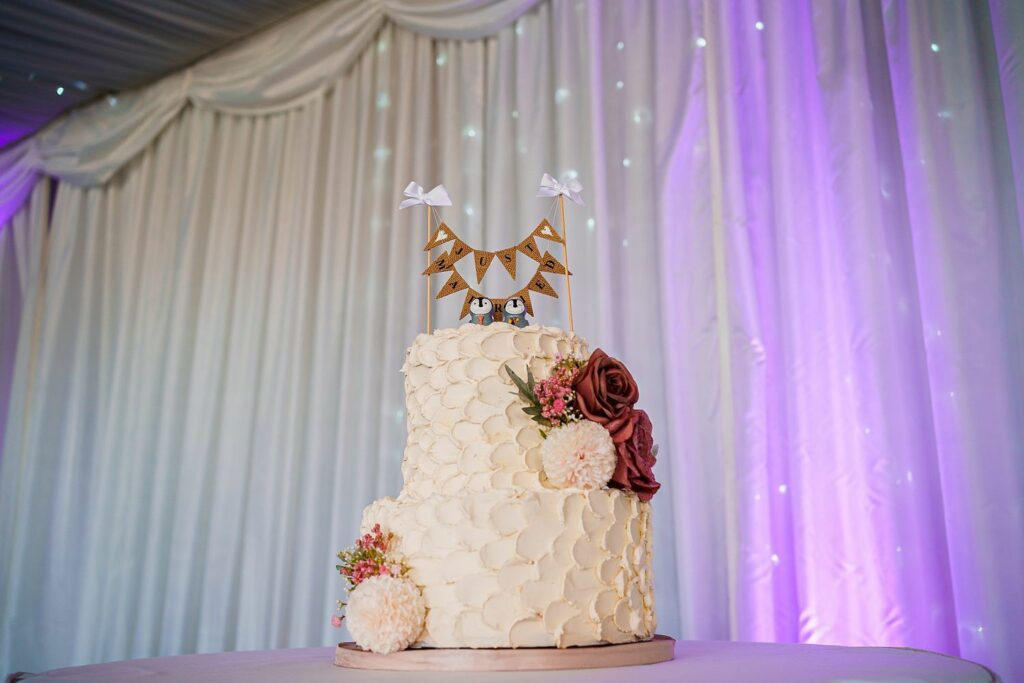 A white, two-tier wedding cake decorated with textured white frosting and adorned with pink flowers and greenery. At the top of the cake, there is a miniature decorative bunting with small white bows. The backdrop features white and lavender drapery. Image by Documentary Wedding Photographer Lewis Bishop Photography.