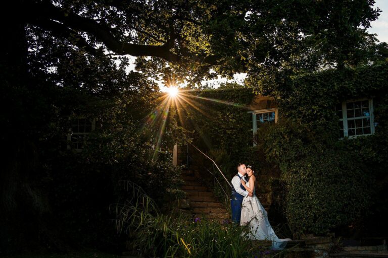 A couple in wedding attire stands closely together, embracing under a large tree with dense foliage. The groom is in a suit with suspenders, and the bride is in a white gown. Sunlight filters through the tree branches, creating a radiant effect in the background. Image by Documentary Wedding Photographer Lewis Bishop Photography.