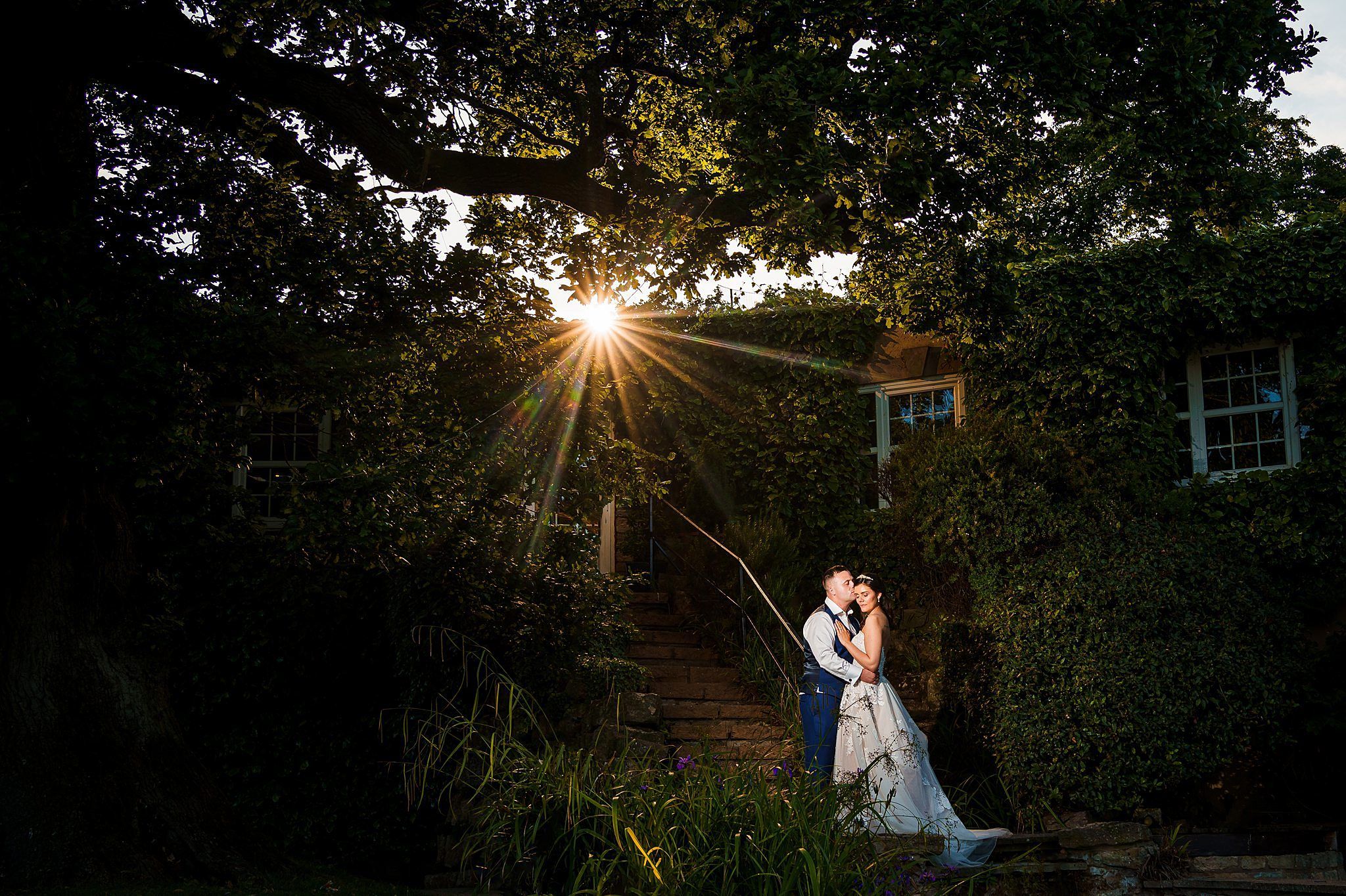 A couple in wedding attire stands closely together, embracing under a large tree with dense foliage. The groom is in a suit with suspenders, and the bride is in a white gown. Sunlight filters through the tree branches, creating a radiant effect in the background. Image by Documentary Wedding Photographer Lewis Bishop Photography.