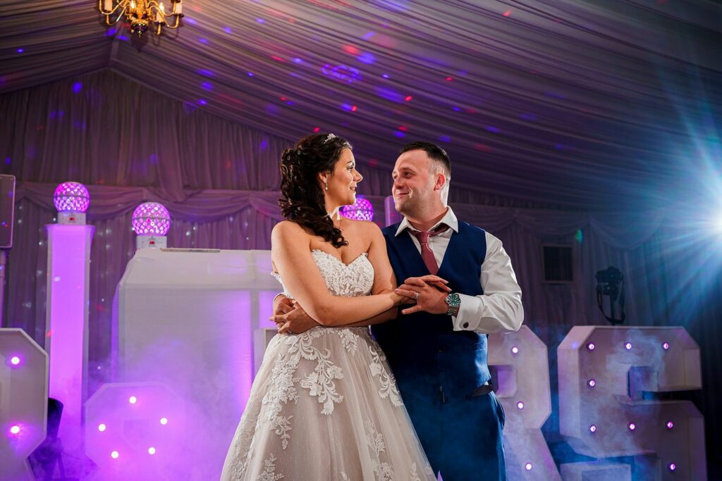A bride in a white lace dress and a groom in a blue waistcoat are dancing and looking at each other under colorful lights in a decorated venue. A chandelier hangs above them, and large illuminated letters are in the background, surrounded by a misty atmosphere. Image by Documentary Wedding Photographer Lewis Bishop Photography.