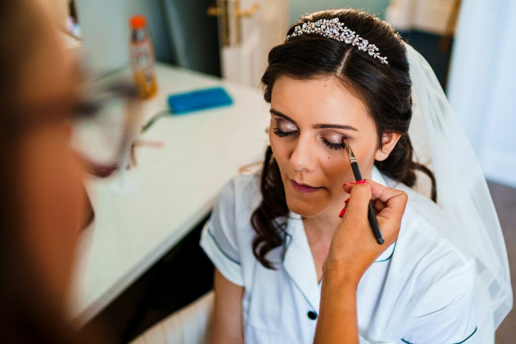 A bride with long dark hair and a jeweled headpiece sits with her eyes closed while a makeup artist applies eyeshadow with a brush. She is wearing a white outfit, and a blurred workspace with makeup accessories is visible in the background. Image by Documentary Wedding Photographer Lewis Bishop Photography.