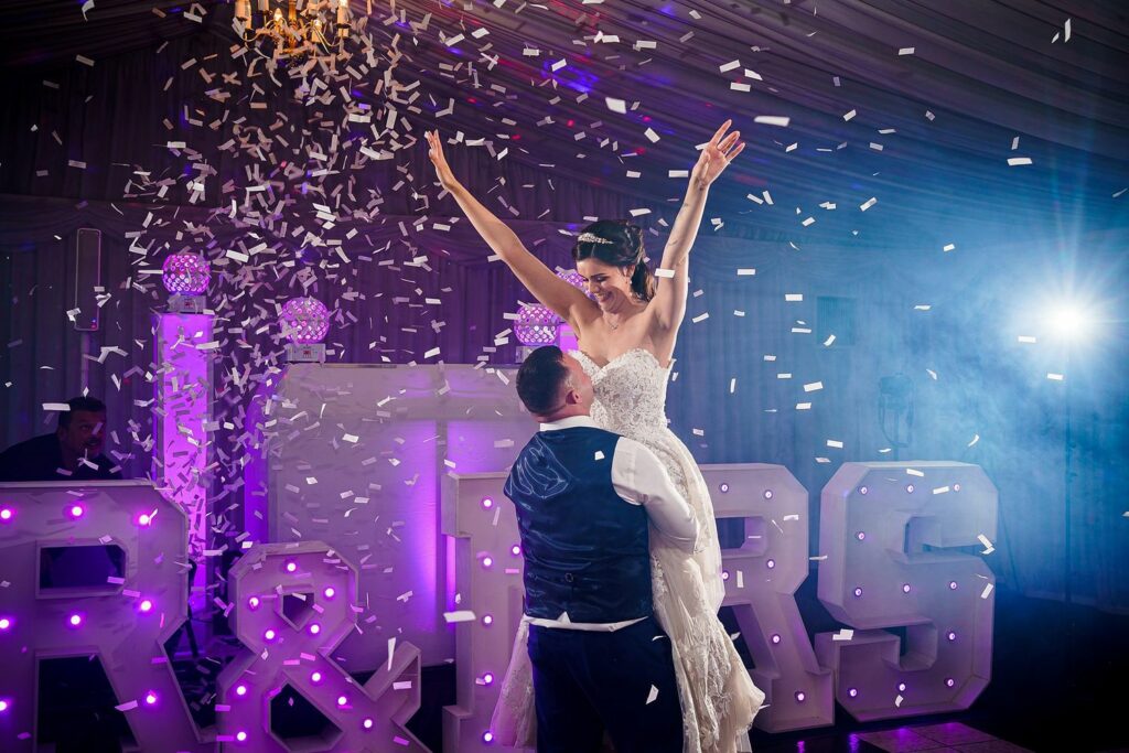 A groom lifts a joyous bride in a strapless wedding dress as white confetti rains down. The background features large, illuminated marquee letters "R & R" and soft, colorful lighting. The scene is lively, celebratory, and filled with festive energy. Image by Documentary Wedding Photographer Lewis Bishop Photography.