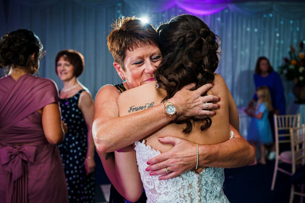 A woman in a black dress with a watch hugs a bride in a white gown. The bride has a tattoo on her shoulder reading "Forever." They are at a celebratory event, with people conversing and children playing in the background. The scene is warmly lit, creating a joyful atmosphere. Image by Documentary Wedding Photographer Lewis Bishop Photography.