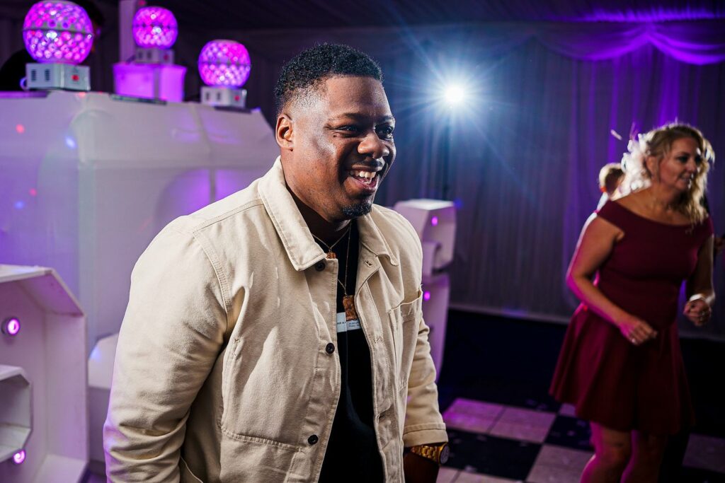 A man in a light-colored jacket, smiling and looking to his right, stands in a dimly lit room with purple lighting. Behind him, a woman in a red dress is dancing, and decorative lights glow in the background. The atmosphere appears cheerful and festive. Image by Documentary Wedding Photographer Lewis Bishop Photography.