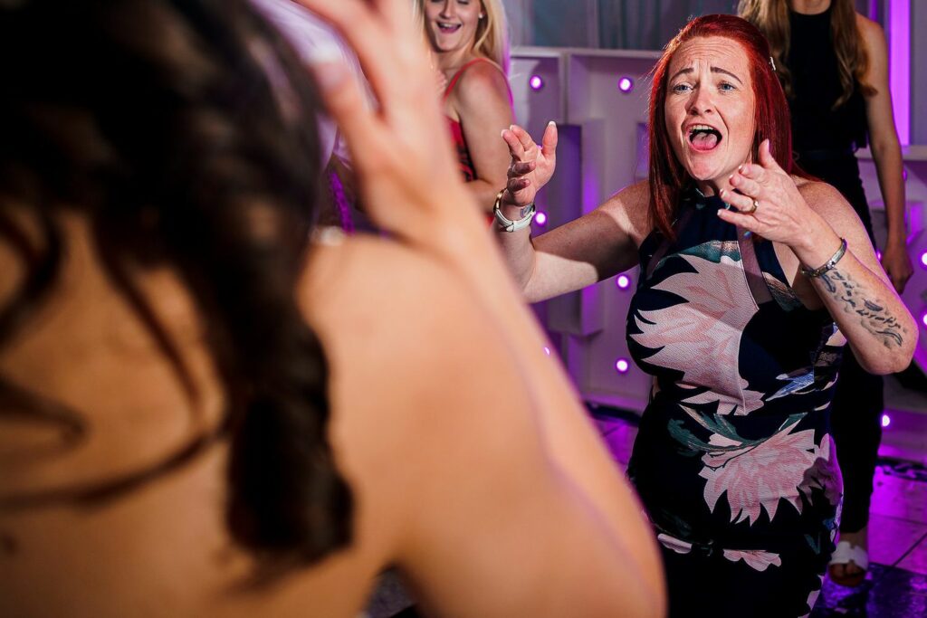 A woman with long red hair and a floral dress joyfully dances with her arms outstretched at an indoor event. Another woman is visible from behind, and people in the background are also engaged in the lively atmosphere. Purple lights adorn the scene. Image by Documentary Wedding Photographer Lewis Bishop Photography.