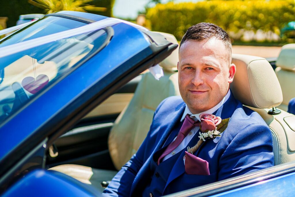 A man in a blue suit with a pink tie and boutonniere sits confidently in the driver's seat of a blue convertible with the top down. The background features greenery and a sunny sky, indicating a pleasant day. A small heart decoration hangs from the rearview mirror. Image by Documentary Wedding Photographer Lewis Bishop Photography.