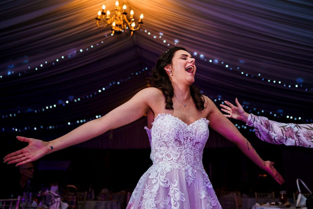 A bride in a white lace gown with her arms outstretched, laughing joyfully under a decorated tent with string lights and a chandelier. The background is softly lit with a festive ambiance, creating a warm and celebratory atmosphere. Image by Documentary Wedding Photographer Lewis Bishop Photography.